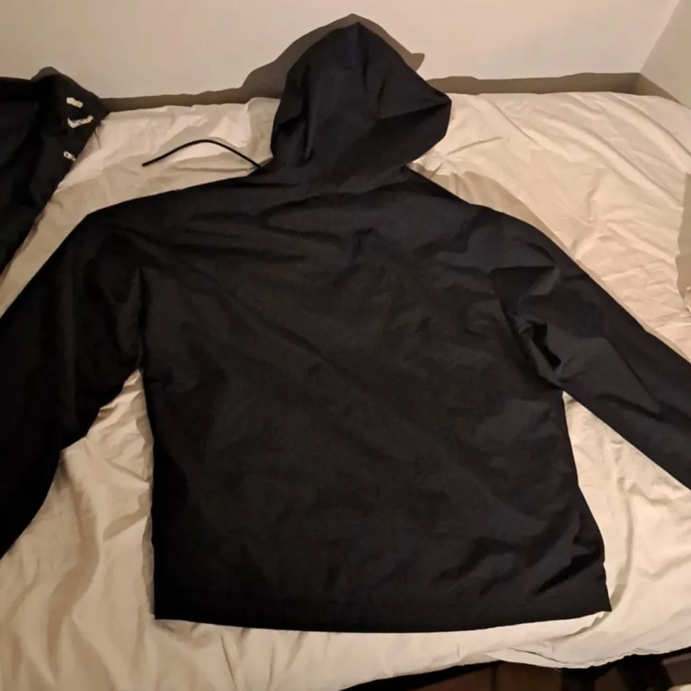 Säljer en svart vindjacka från Under Armour med grått bröstparti och logga. Jackan har huva med dragsko och dragkedja framtill. Perfekt för träning eller när det blåser ute.. Takit.