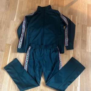 Grön tracksuit från Daily Paper - Säljer ett par svarta träningsbyxor från Daily Paper med resår i midjan och vita/röda sidoremsor med logotyp. Byxorna har en sportig look och raka ben, perfekta för en avslappnad stil.