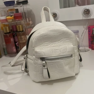 Vit miniryggsäck med dragkedja - Säljer en liten vit ryggsäck i syntetmaterial med silverfärgade detaljer och flera dragkedjefack. Väskan har justerbara axelremmar och en liten ficka framtill. Perfekt för att bära det viktigaste på stan eller till skolan.