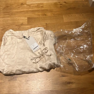 Ljus krämfärgad långklänning från ÀVORA - Säljer en elegant axelbandslös långklänning från ÀVORA i ljus krämfärgad satin. Klänningen har smock upptill och en mjuk, följsam passform. Perfekt för sommarens tillfällen. Märke och tagg kvar. Tyvärr passar den inte mig.