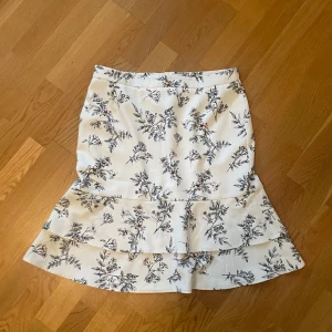 Vit blommig kjol från MQ - Säljer en vit kjol från MQ med svart blommigt mönster. Kjolen har volangdetaljer och är i en kort modell. Perfekt för dig som gillar romantisk stil och vill ha något som sticker ut lite extra. Perfekt för sommardagar! Nypris=600kr