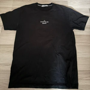 Svart t-shirt från Stone Island - Säljer en svart t-shirt från Stone Island med liten vit logga framtill och tryck på ryggen. T-shirten har klassisk passform och är tillverkad i mjuk bomull. Perfekt för dig som gillar streetwear och vill ha något stilrent med coola detaljer. Den är storlek XL men passar bra som en L med 