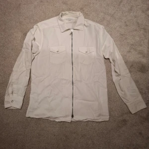 Vit overshirt med dragkedja från Eton - Snygg vit overshirt från Eton med dragkedja framtill och två bröstfickor med knapp. Skjortan har klassisk krage och långa ärmar med manschett. Perfekt att slänga på över en t-shirt för en clean look. Nypris 1400 kr