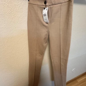 Beige kostymbyxor från MNG, stl 34 - Snygga beige kostymbyxor från MNG i storlek 34. Byxorna har hög midja, raka ben och stängs med knapp och dragkedja. Perfekta för en clean och stilren look. Materialet är mjukt och känns bekvämt mot huden.