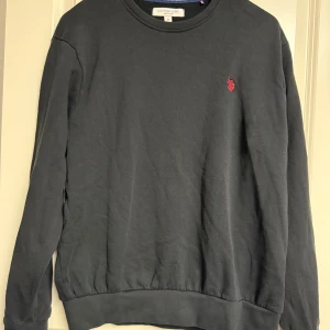 Svart sweatshirt från U.S. Polo Assn. - En svart sweatshirt från U.S. Polo Assn. med rund hals och diskret röd logga broderad på bröstet. Tröjan har långa ärmar och ribbade muddar vid ärmslut och nederkant. Perfekt för en chill och stilren look.