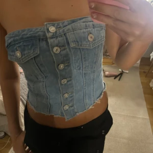 Ljusblå jeans bandeau tubtopp - Säljer en unik ljusblå bandeau tubtopp i jeans med snygga silverfärgade knappar framtill och rå, fransig kant nedtill. Toppen har dekorativa bröstfickor och cool denimlook som sticker ut. Perfekt för dig som vill ha något edgy och trendigt.