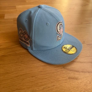 Ljusblå New Era 59FIFTY keps Mariners - Snygg ljusblå 59FIFTY keps från New Era med Seattle Mariners-logga framtill och 20th Anniversary patch på sidan. Klassisk flat brim och broderad MLB-logga bak. Tillverkad i 100% polyester, perfekt för dig som gillar sportig streetstyle.