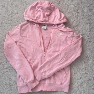 Rosa hoodie från Nike med dragkedja - Säljer en ljusrosa hoodie från Nike med huva och hel dragkedja framtill. Tröjan har långa ärmar, två fickor och en broderad vit Nike-logga på bröstet. 