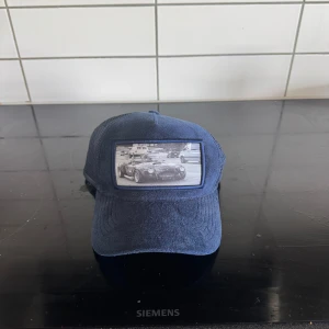 Blå keps från Emilio Pendenza med bilmotiv - Snygg blå keps från Polo Pericoli med mesh på sidorna och en cool patch framtill med svartvit bild på en sportbil. Kepsen har böjd skärm och justerbar baksida. Perfekt för dig som gillar bilar och streetstyle.
