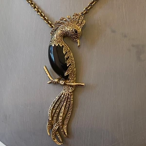 Guldigt halsband med fågelhänge - Unikt halsband med en stor, detaljerad fågel i guldtonad metall och svart emalj på kroppen. Kedjan är grov och har en vintagekänsla. Fågeln har snygga detaljer på fjädrarna och sitter på en gren. Perfekt statement-piece för dig som vill sticka ut.