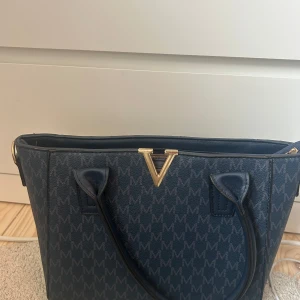 Mörkblå handväska med gulddetaljer  - Snygg blå handväska med guld detaljer och med guldigt V-spänne och mönster av bokstaven M över hela väskan. Väskan har två handtag, dragkedja upptill och ljus insida med innerfack med dragkedja. Lite smutsig inuti men går att få bort enkelt.