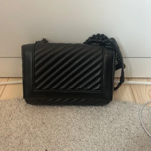 Svart quiltad axelväska med kedja - Snygg svart axelväska i quiltad skinnimitation med diagonala ränder. Väskan är smutsig inuti men går att få bort.