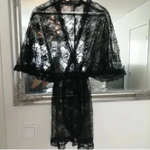 Spetskimono med knytband - Snygg kort svart spetskimono med omlottmodell och knytband i midjan. Klänningen har vida, korta ärmar och vacker blommig spets över hela plagget. Använts 1-2ggr, etikett bortklippt 