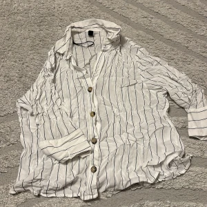 Vit randig skjorta med knappar - Vit skjorta med svarta tunna ränder och bruna knappar framtill. Skjortan har lång ärm som kan vikas upp och en avslappnad passform. Perfekt för en stilren och enkel look. Tillverkad i ett lätt och luftigt material.