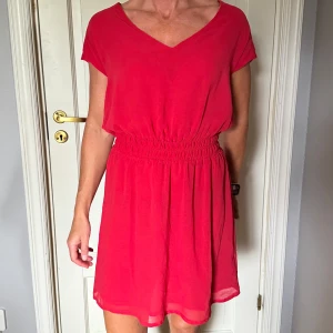 Röd klänning med v-ringning - Superfin röd kortklänning med v-ringad hals och resår i midjan som ger en snygg siluett. Klänningen har korta ärmar och är gjord i ett lätt och luftigt material, perfekt för varma dagar.