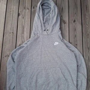 Grå Nike hoodie med huva, stl S - Klassisk grå hoodie från Nike i storlek S men det står M. Tröjan har en stor huva, känguruficka framtill och Nike-logga broderad på bröstet. Tillverkad i mjuk bomullsblandning, perfekt för chill dagar eller när du vill ha en enkel streetstyle-look.