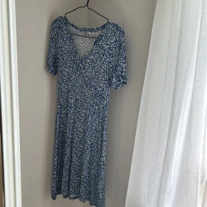 Blå blommig omlottklänning - Supersöt blå klänning med vitt blommönster och omlott-v-ringning. Klänningen har korta ärmar och faller till knäna. Perfekt för varma dagar och har en luftig, skön passform.
