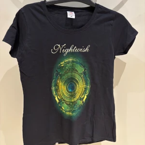 Nightwish t-shirt från Fruit of the Loom - Svart t-shirt med Nightwish-tryck fram och bak i grönt och gult. Modellen är Lady Fit med rund hals och korta ärmar. Materialet är mjuk bomull och passformen är normal. Perfekt för dig som älskar bandmerch och vill sticka ut med snygg grafik.