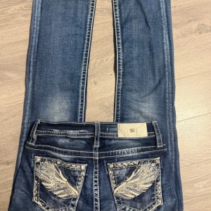 Miss Me jeans med broderade vingar fickor - Snygga blå jeans från Miss Me med broderade bakfickor och paljettdetaljer. Jeansen har kontrastsömmar, klassisk femficksmodell och en cool tvättad look. Perfekta för dig som gillar detaljer och vill sticka ut lite extra.