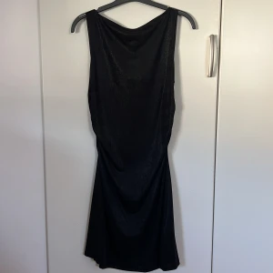 Svart kortklänning - Snygg svart kortklänning med scrunch på sidorna. Klänningen har ärmlös design och en lätt figurnära passform som blir utsvängd vid kjolen. Materialet är mjukt och följsamt med metallic-effek. Aldrig använd