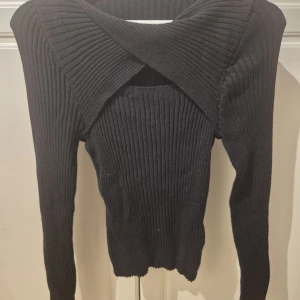 Svart ribbad stickad tröja omlott - Säljer en svart ribbstickad tröja med lång ärm och snygg omlottdetalj vid halsen. Tröjan är figurnära och har en cool, modern look som passar perfekt till jeans eller kjol. Materialet är mjukt och stretchigt för extra komfort.
