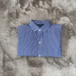 Blåvit randig skjorta från Ralph Lauren - Klassisk blå och vit randig skjorta från Ralph Lauren med broderad logga på bröstet. Skjortan har lång ärm, knappar framtill och krage. Tillverkad i mjuk bomull och passar perfekt till en stilren look.