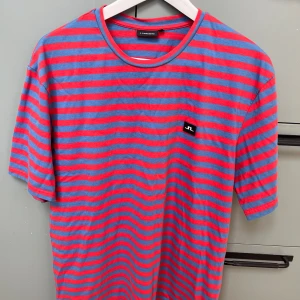 Randig t-shirt från J.Lindeberg - Snygg t-shirt från J.Lindeberg med blå och röda horisontella ränder. Klassisk passform med rund hals och korta ärmar. Liten svart JL-logga på bröstet. Perfekt för dig som gillar färg och stilren design. Ej byte/öppet köp. 