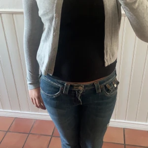 Grå kofta från 157, storlek S - En enkel och stilren grå kofta från 157 i storlek S. Koftan har rund hals, långa ärmar och knäppning med matchande knappar framtill. Materialet är mjukt och stickat, perfekt att slänga över en t-shirt eller linne. Passar dig som gillar basic och clean look. Har en liten blå fläck längst ut på ärmen