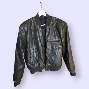 Skinnjacka i konstläder i storlek L från Divided - Snygg jacka med dragkedja och fickor framtill. Jackan har muddar vid ärmslut och nederkant, samt en liten ficka med dragkedja på bröstet. Den har också mindre axelvaddar.