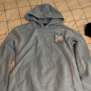 Ljusblå hoodie från Amiri - Säljer en ljusblå hoodie från Amiri med vit logga på bröstet och stort AMIRI-tryck på ryggen. Tröjan har huva, känguruficka och är tillverkad i mjukt material. Perfekt för dig som gillar streetwear och vill sticka ut med en snygg färg.
