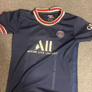 PSG Messi #30 marinblå matchtröja - Snygg PSG fotbollströja i marinblått med Messi och nummer 30 på ryggen. Tröjan har korta ärmar, klubbmärke på bröstet och sponsortryck framtill. Kragen och ärmsluten har röd, vit och blå randig detalj. Tillverkad i polyester, perfekt för fotbollsfans.