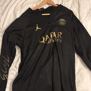 Svart PSG långärmad fotbollströja - Snygg svart långärmad PSG-tröja från Jordan med gulddetaljer och Qatar Airways-tryck på bröstet. Paris-logga på ärmen och klubbmärke på bröstet. Tillverkad i lätt och ventilerande material, perfekt för träning eller match.