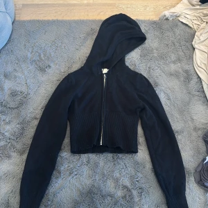 Svart croppad stickad hoodie med dragkedja - Svart croppad hoodie i stickat material med ribbad midja och långa ärmar. Tröjan har huva och hel dragkedja framtill, vilket ger en chill och trendig vibe. Perfekt att slänga på sig över ett linne eller t-shirt.