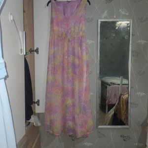 Pastellfärgad långklänning med v-ringning - Långklänning i mjuka pastellfärger med abstrakt mönster i lila, rosa och gul. Klänningen har v-ringning och är ärmlös, med ett lätt och luftigt tyg som ger en flowy känsla. Perfekt för varma dagar och somriga vibes.
