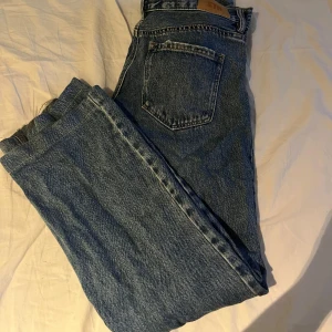 Strandvarius  midwaist blå jeans straight fit - Klassiska jeans i blå denim med rak passform och femficksdesign. Jeansen har knappgylf och tydliga sömmar, samt en något ljusare tvätt. Perfekta för dig som gillar tidlös stil och vill ha ett par riktigt schyssta jeans i garderoben. Lite slitna på ena benet 