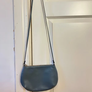 Blå axelväska från H&M i skinnimitation - Stilren blå axelväska från H&M i skinnimitation med rundad form och guldfärgad dragkedja. Väskan har en smal axelrem och minimalistisk design, perfekt för att bära det viktigaste. Enkel och snygg att matcha till olika outfits.