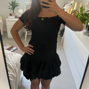 Svart offshoulder kortklänning - Supersöt svart kortklänning med offshoulder och smockad överdel. Kjolen är puffig där nere men sitter närmare kroppen på överkroppen. Perfekt för dig som gillar en feminin och trendig stil. Storlek M men passar XS-M.
