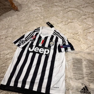 Juventus Pogba 10 Adidas matchtröja M - Juventus hemmatröja med Pogba 10 på ryggen, tillverkad av Adidas i ClimaCool-material. Klassisk svartvit randig design, kortärmad med officiella Champions League och UEFA Respect-patchar. Jeep-logga på bröstet och broderade klubbmärken framtill.