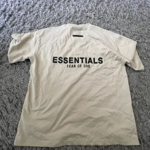Beige Essentials Fear of God t-shirt - Snygg beige t-shirt från Essentials Fear of God med svart logga tryckt på ryggen. Klassisk passform med rund hals och korta ärmar. Perfekt för en clean och stilren stil. (Samma som marlon använder) Tillverkad i mjuk bomull som känns skön mot huden. Pris lapp finns kvar här aldrig använt 