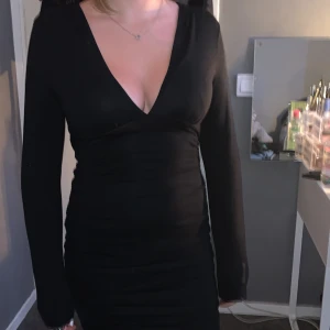 Svart tight klänning från Amisu - Säljer en svart, tight kortklänning från Amisu med lång ärm och djup v-ringning. Klänningen har rynkade detaljer längs sidorna och är figurnära med stretchigt material. Perfekt för dig som gillar en stilren och enkel look.