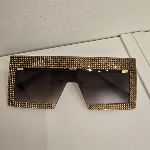 Guldglittriga oversized solglasögon - Statement solglasögon med stora, rektangulära linser och en ram täckt av glittrande guldiga stenar. Snygg futuristisk vibe och mörka tonade glas. Perfekta för dig som vill sticka ut och glänsa extra mycket.