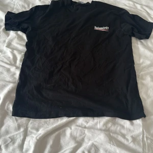 Svart Balenciaga t-shirt i bomull - Svart t-shirt från Balenciaga i mjuk bomull med klassisk passform. Tryckt Balenciaga-logga i vitt och rött på bröstet. Enkel och clean design med rund hals och korta ärmar. Perfekt för dig som gillar streetstyle och vill ha något från ett ikoniskt märke.