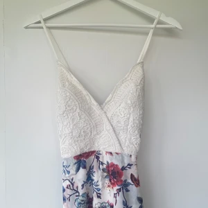 Vit spetsklänning med blommigt mönster - Långklänning med vit spets upptill och smala axelband. Kjolen är vit med blommigt mönster i blått och rött. V-ringad omlott upptill och dragkedja i sidan. Perfekt för sommarens alla tillfällen.
