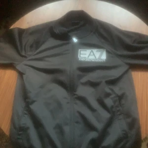 Svart jacka från Emporio Armani EA7 - Säljer en svart jacka från Emporio Armani EA7 med stor logga på bröstet. Jackan har dragkedja framtill, ståkrage och två fickor. Perfekt för dig som vill ha en stilren och sportig look. 