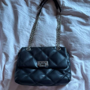 Svart quiltad axelväska med kedja - Snygg svart axelväska i quiltad skinnimitation med silverfärgad kedjerem och vridlås framtill. Väskan har ett innerfack med dragkedja och detaljer i metall. Perfekt storlek för det viktigaste och ger en cool vibe till din outfit. Är i fint skick, skriv för fler frågor💗💗 pris kan diskuteras 💗💗
