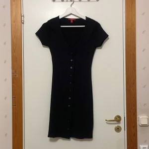 Svart ribbad kortklänning från Guess - Supersnygg svart kortklänning från Guess med ribbad struktur, v-ringning och knappar framtill. Klänningen har korta ärmar och en figurnära passform. Perfekt för dig som gillar stilrena och trendiga plagg med diskret logga nertill. Använd endast 1 gång!