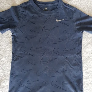 Blå Nike t-shirt med logomönster - Snygg blå t-shirt från Nike med korta ärmar och all-over print av svarta Nike Swoosh-loggor. Klassisk rund halsringning och mjukt bomullsmaterial. Perfekt för dig som gillar sportig och avslappnad stil.