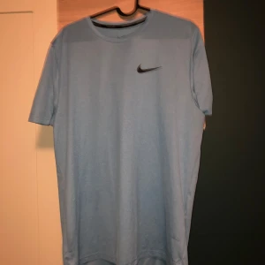 Blå Nike Dri-Fit t-shirt - Säljer en blå Nike Dri-Fit t-shirt med klassisk svart swoosh-logga på bröstet. T-shirten är i sportig modell med rund hals och korta ärmar, tillverkad i ett lätt och snabbtorkande material som passar perfekt för träning.