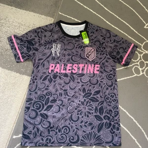 FC Palestina blommig fotbollströja M - Unik fotbollströja från FC Palestina i mörkgrått med blommigt mönster och rosa detaljer. Tryck med texten PALESTINE och klubbmärke på bröstet, samt rosa grafik på ryggen. Tillverkad i lätt och ventilerande material, perfekt för match eller träning.