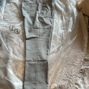 Grå rutiga kostymbyxor herr 44 - Snygga grå kostymbyxor med diskret rutigt mönster. Klassisk passform med fickor bak och hällor för bälte. Materialmix av polyester, viskos och elastan för skön stretch. Perfekta för en clean och stilren look.
